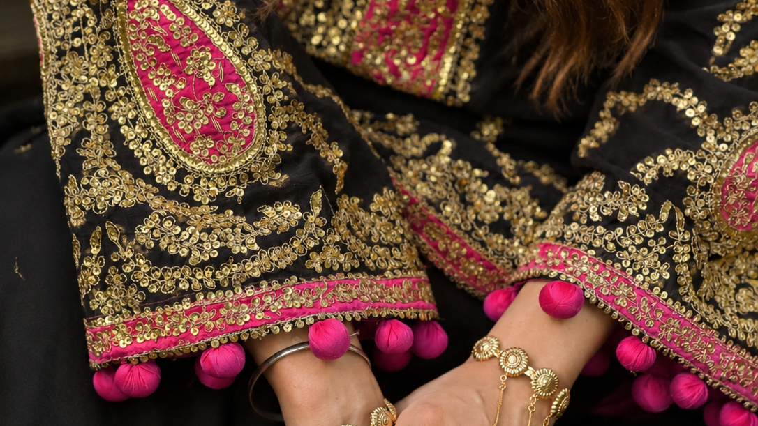 Close-up of a person wearing an ornate black traditional outfit with heavy gold embroidery and bright pink accents, along with matching gold jewelry and bangles.
